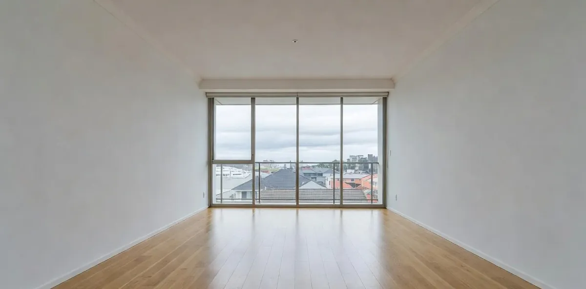 An empty open-plan apartment floor running corner to corner, balcony glass and rooftops beyond